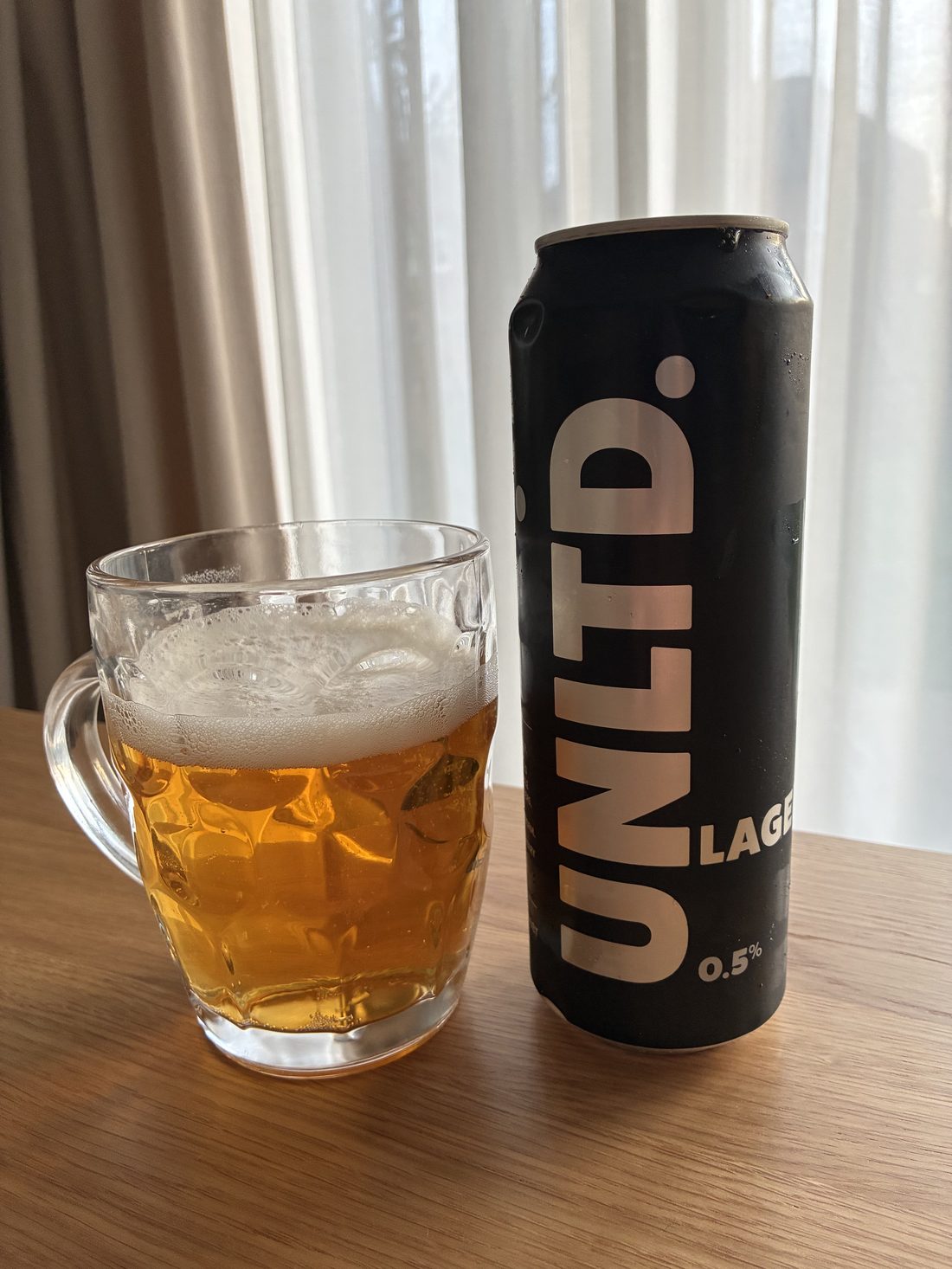 A pint of UNLTD alcohol-free lager poured into a dimpled mug beside the can &mdash; pale golden and clear with a light head