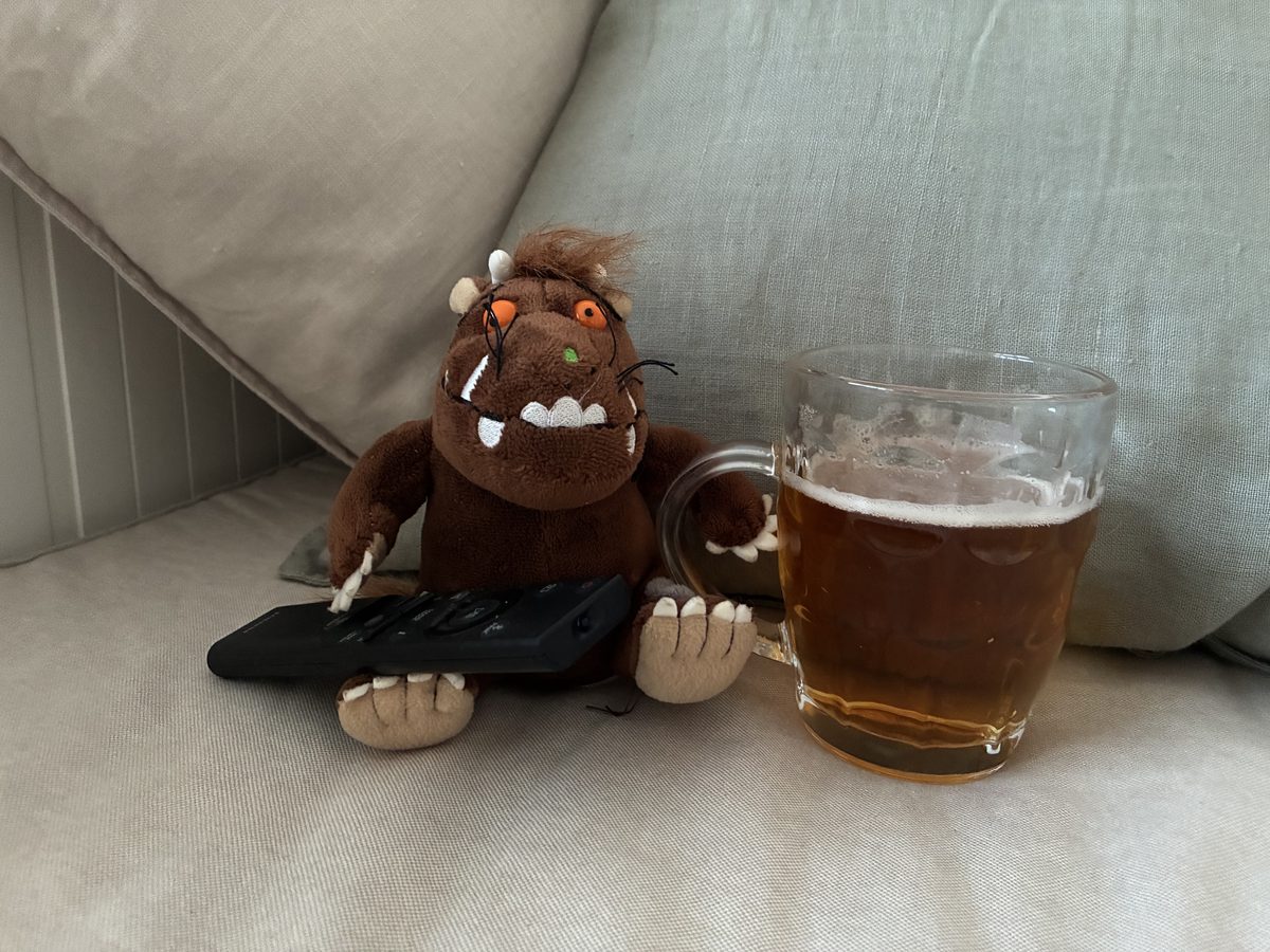 A cuddly monster toy holding a TV remote beside a pint of UNLTD alcohol-free beer on the sofa